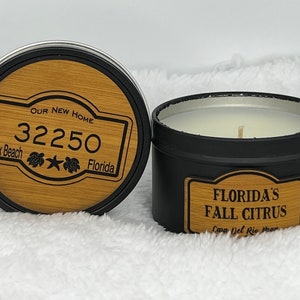 May include: Two black candle tins. One tin has a wooden lid with the text "Our New Home 32250 Jax Beach Florida" and a design of two sea turtles and a star. The other tin has a label that reads "Florida's Fall Citrus Casa Del Rio Manor".