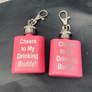 Op de afbeelding: Twee roze heupflacons met zilveren accenten en bevestigde clips. De heupflacons hebben de tekst "Cheers to My Drinking Buddy!" in zilveren letters. De heupflacons zijn rechthoekig en lijken van metaal te zijn.