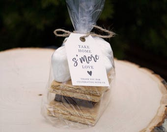 Op de afbeelding: Een doorzichtige plastic zak gevuld met een s'mores-set. De set bevat twee graham crackers, een chocolade reep en twee marshmallows. Een wit label met zwarte tekst luidt "Take home s'more love" met een klein hartpictogram. Het label zegt ook "Thank you for celebrating with us".
