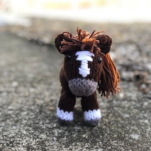 Puede incluir: Un caballo de juguete de punto marrón con marcas blancas en la cara y las patas. El caballo tiene una larga melena y cola marrones.