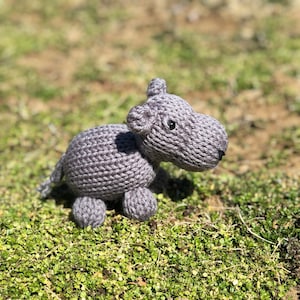 Puede incluir: Un animal de peluche gris tejido que se asemeja a un hipopótamo, sentado en un lecho de musgo verde.