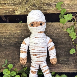 May include: A white crocheted mummy doll with brown yarn for the body and black eyes. The doll is standing on a bed of green leaves.