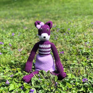 May include: A purple knitted toy animal with white stripes and a pink bow sitting in a field of purple flowers.