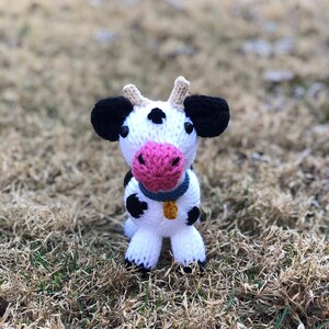 Puede incluir: Un juguete de vaca de punto blanco y negro con orejas rosas y un collar azul. El juguete está de pie sobre un lecho de hierba marrón.