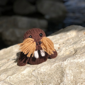 May include: A brown knitted walrus plush toy with a tan yarn moustache and white yarn tusks. The toy is sitting on a light brown rock.