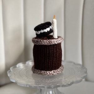 May include: A brown knitted cake with a white and black Oreo cookie on top and a white candle. The cake is on a clear glass cake stand.