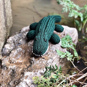 Puede incluir: Un juguete de cocodrilo de punto verde sentado en una roca con follaje verde en el fondo.