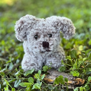 Puede incluir: Un pequeño juguete de perro de crochet, gris y esponjoso, con ojos marrones y una nariz negra. El juguete está sentado en una cama de hojas verdes.
