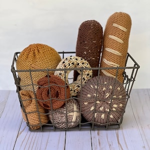 Puede incluir: Una cesta de alambre metálico llena de varios artículos de comida de punto, incluyendo una hogaza de pan, una baguette, una dona, un pretzel y una hogaza de pan redonda. La cesta está sentada sobre una superficie de madera clara.