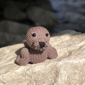 Puede incluir: Un juguete de foca tejido a mano de color marrón, con ojos negros y nariz negra. La foca está tumbada sobre una roca clara. El juguete tiene un diseño sencillo y está hecho de hilo suave.