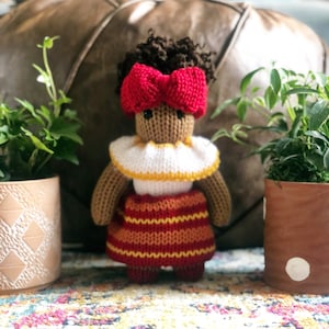 Puede incluir: Una muñeca de punto con piel marrón, cabello castaño rizado y un lazo rojo. La muñeca lleva una camisa de cuello blanco y amarillo y una falda de rayas rojas y amarillas. La muñeca está de pie sobre una alfombra colorida.