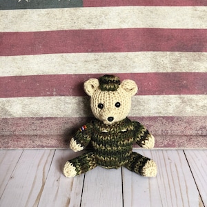 May include: A crocheted teddy bear wearing a camouflage uniform and a hat. The bear is sitting in front of an American flag.