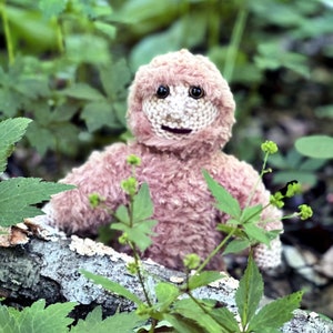 May include: A pink crocheted plush toy resembling a creature with a furry face and large eyes. The toy is sitting in a bed of green leaves.