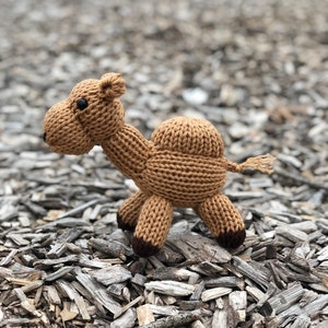 Puede incluir: Un juguete de camello de punto marrón con ojos negros. El camello está de pie sobre una cama de virutas de madera.