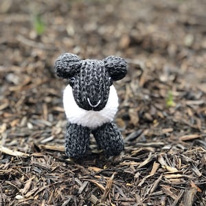 May include: A black and white knitted sheep toy with a white fluffy face. The toy is sitting on a bed of brown wood chips.