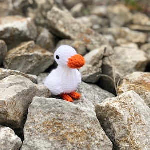Puede incluir: Un pájaro blanco de crochet con pico y patas naranjas sentado en una pila de rocas grises.