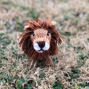 Puede incluir: Un juguete de león de punto marrón y naranja con rasgos faciales blancos. El juguete está sentado en un parche de hierba verde.