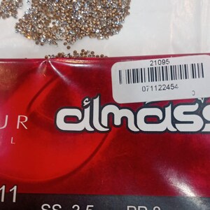 May include: A collection of small, sparkling crystals in shades of gold and silver. The crystals are scattered on a white surface, with a red and black label visible. The label includes the text "SS 3.5 Crystal 10 Gross".