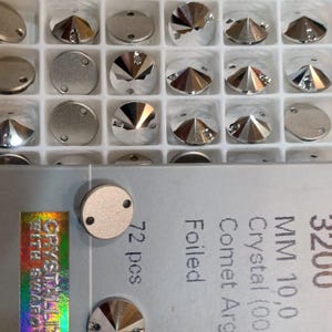Op de afbeelding: Een verzameling zilverkleurige kristallen strass-steentjes in een witte tray. Sommige zijn plat en rond, andere gefacetteerd en puntig. De kristallen zijn 10,0 mm groot en van het type "Comet Argent Light".