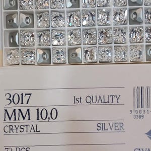 Puede incluir: Una caja de pedrería de cristal plateado, cada una de aproximadamente 10 mm, dispuestas en una cuadrícula. La caja incluye el texto "3017 MM 10,0 CRYSTAL SILVER 72 PCS. Made in Austria."