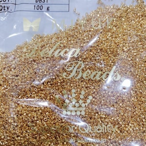 Peut inclure: Un sac en plastique transparent rempli de petites perles Delica dorées. Le sac porte une étiquette indiquant "Delica Beads", taille 11/0, couleur DB31 et une quantité de 100 g. Les mots "Delica Beads" sont également imprimés sur le sac.