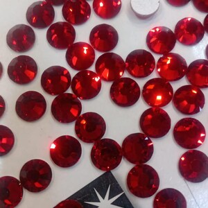 Può includere: Una collezione di cristalli rotondi, sfaccettati, rosso rubino, sparsi su una superficie bianca. I cristalli sono altamente riflettenti, catturando la luce e creando un effetto scintillante. Sono presenti anche un piccolo adesivo argentato e un'etichetta nera con la scritta "GENUINE CZECH CRYSTAL".