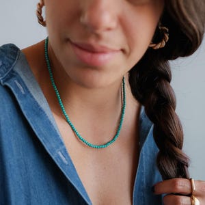 May include: A delicate necklace featuring small, round, vibrant turquoise-colored beads. The necklace is worn with gold-toned twisted hoop earrings and a gold-toned ring, paired with a blue denim shirt. The jewelry pieces are simple and minimalist in their design, highlighting the bright color of the beads.