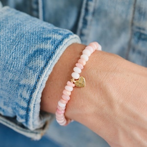 Peut inclure: Bracelet de perles roses et blanches avec un charm en forme de cœur doré. Le bracelet est composé de petites perles rondes roses et blanches. Un charm en forme de cœur doré est suspendu au centre. Le bracelet est porté au poignet.
