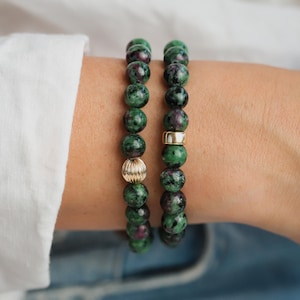 May include: Two beaded bracelets. Each bracelet features green and maroon gemstone beads. One bracelet has a gold-colored, ribbed bead, and the other has a gold-colored, smooth cylindrical bead. The bracelets are worn on a wrist.