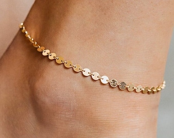 May include: A delicate gold-colored anklet featuring small, round, flat disc charms. The anklet is shown worn on an ankle, with the skin tone visible. The jewelry is simple and elegant.