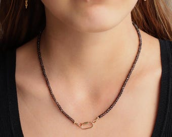 May include: A necklace featuring small, faceted dark gray beads strung together, finished with a gold-toned oval carabiner-style clasp positioned at the front. The beads have a reflective, shimmering quality, and the metal clasp provides a contrast.