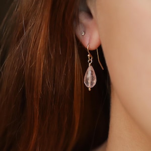 May include: A gold earring with a pear-shaped, pale pink gemstone. The earring is suspended from a pierced ear. The background features reddish-brown hair and a glimpse of a person's shoulder.