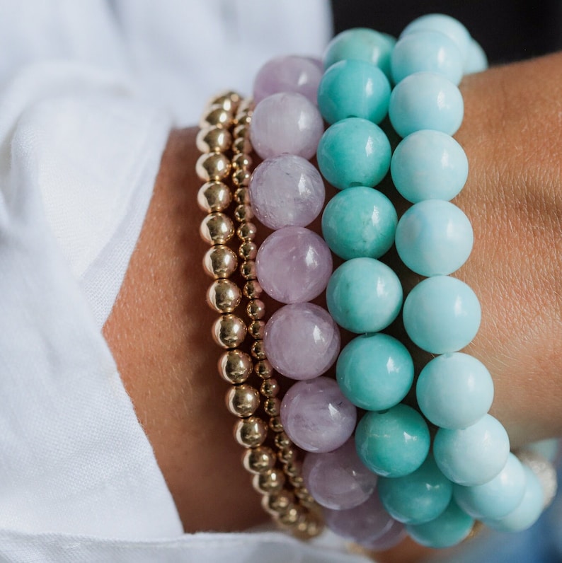May include: A selection of beaded bracelets in a close-up shot. The bracelets feature gold-coloured beads, lilac-coloured beads, and turquoise and pale blue beads. The bracelets are stacked, creating a layered effect.