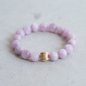 May include: A bracelet with pale lilac-coloured round beads and a central gold-coloured, ribbed bead. The bracelet is presented on a light grey surface. The beads are approximately 1 cm in diameter.