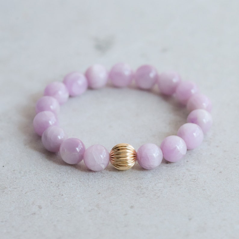 May include: A bracelet with pale lilac-coloured round beads and a central gold-coloured, ribbed bead. The bracelet is presented on a light grey surface. The beads are approximately 1 cm in diameter.