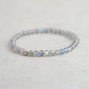 May include: A delicate bracelet featuring small, round, gray beads with iridescent blue and yellow flashes. A single gold-colored bead is incorporated into the design. The bracelet is likely made of gemstone beads and is designed to be worn on the wrist.