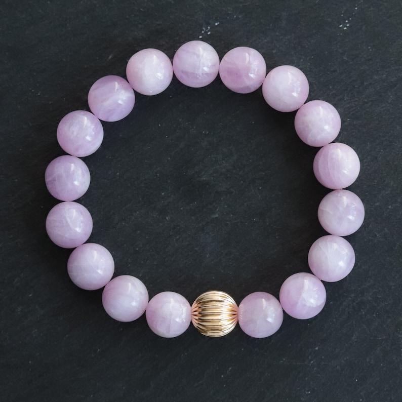 May include: A bracelet made of pale lavender, spherical beads, with a single gold-coloured, ridged bead. The jewellery is presented on a dark, textured surface. The beads are approximately 1.5 cm in diameter.