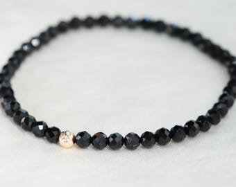 May include: A delicate bracelet composed of small, faceted black beads arranged in a circular pattern, featuring a single, textured, metallic rose gold-toned bead positioned as a focal point within the continuous strand of dark stones.