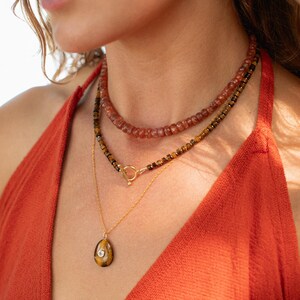 May include: A layered necklace set consisting of a top strand of reddish-brown faceted beads, a middle strand of dark brown and gold-toned beads with a gold-toned toggle clasp, and a bottom gold-toned chain featuring a teardrop-shaped tiger eye pendant.