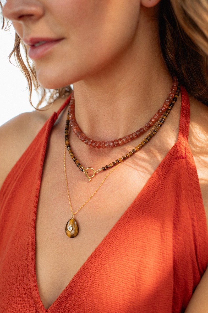 May include: A layered necklace set consisting of a top strand of reddish-brown faceted beads, a middle strand of dark brown and gold-toned beads with a gold-toned toggle clasp, and a bottom gold-toned chain featuring a teardrop-shaped tiger eye pendant.