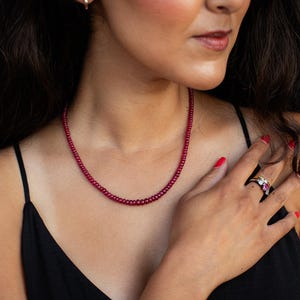May include: A red beaded necklace and a ring with a red gemstone are displayed. The necklace is composed of small, round beads. The ring is on a hand with red nail polish. The model is wearing a black top.