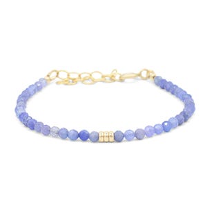 May include: A delicate bracelet featuring small, faceted, light blue tanzanite beads strung together. The center of the bracelet is accented with three smooth, cylindrical gold beads, and it includes a gold-toned metal chain extender.