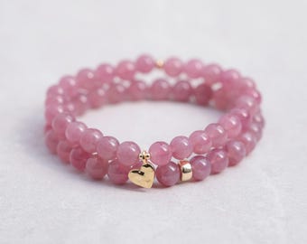 May include: Two pink beaded bracelets. Each bracelet is made of round, light pink beads. One bracelet has a small gold heart charm, and the other has a small gold bead. The bracelets are stacked on top of each other.