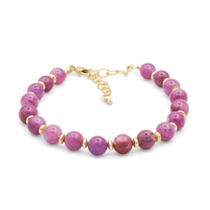 May include: A beaded bracelet featuring round, polished magenta-coloured stones alternating with small, smooth gold-toned metal spacers. The bracelet includes a gold-toned lobster claw clasp and an adjustable chain link extender for sizing.