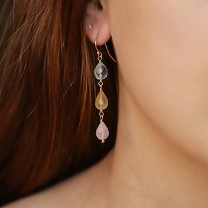 May include: Gold-tone drop earrings with three teardrop gemstones in light blue, yellow, and pink. The gemstones are suspended from a delicate gold wire, creating a layered effect. The earrings are designed to dangle gracefully.