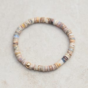 May include: A beaded bracelet featuring a mix of neutral-toned, small, flat beads in shades of beige, tan, and pale pink. A single gold-colored bead is visible. The bracelet is circular and appears to be strung on an elastic cord.