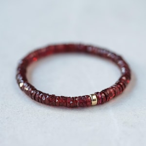 May include: A bracelet made of small, faceted, deep red gemstones. The bracelet is circular with a small gold-coloured accent bead. The gemstones are tightly strung together, forming a continuous band of colour.