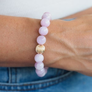 May include: A bracelet with pale pink, round beads and a gold-coloured, textured central bead. The bracelet is displayed on a wrist, with a white top and denim jeans visible in the background.