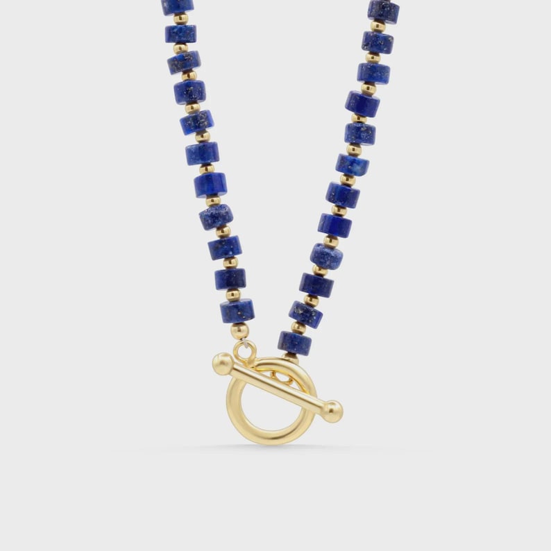 May include: A necklace featuring a repeating pattern of cylindrical deep blue lapis lazuli beads and small gold-toned spherical spacers. The piece is finished with a gold-toned toggle clasp consisting of a circular ring and a horizontal bar.