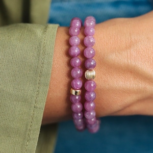 May include: Two beaded bracelets featuring round, polished purple gemstone beads. One bracelet includes a smooth, cylindrical gold-toned accent bead, while the other features a ribbed, spherical gold-toned bead positioned among the purple stones.